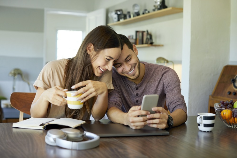 Cheerful boyfriend showing mobile phone to girlfriend in dining room. Young woman is holding coffee cup. They are spending leisure time together at home.