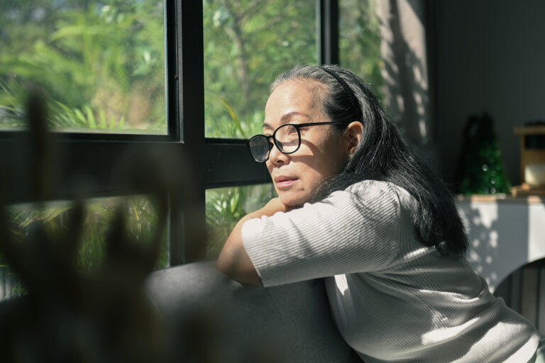 Woman of Asian heritage sits on a couch, leaning against the back and looking out a window thoughtfully