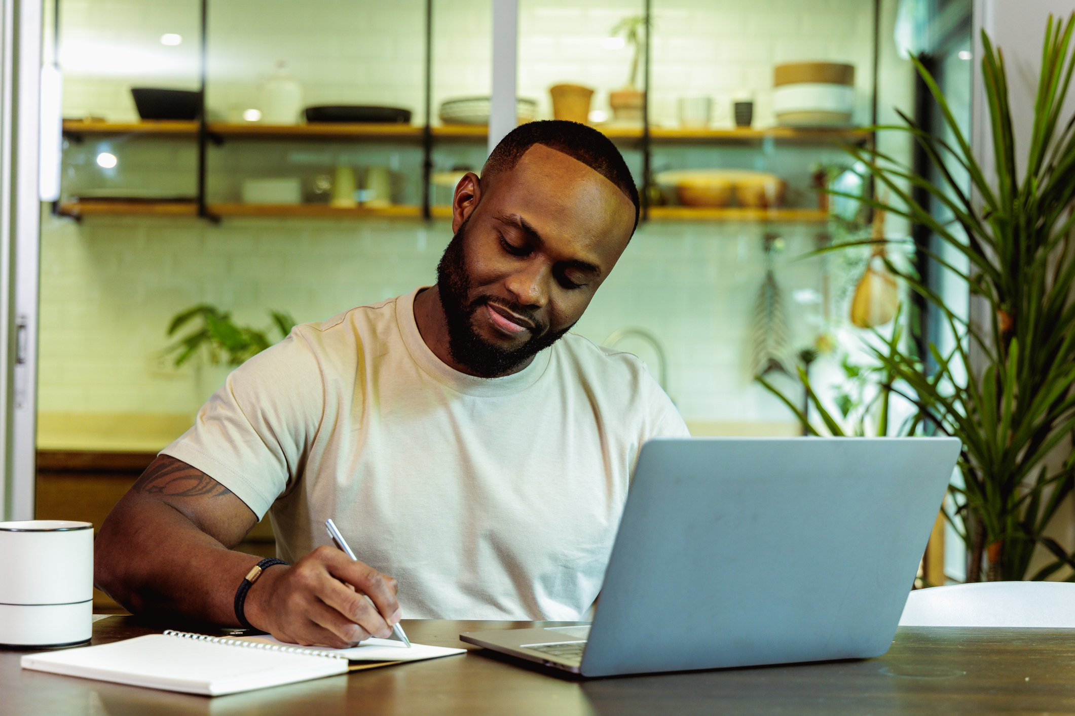 A Black man sits at a table in front of a laptop and takes notes on paper